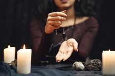 Eine Frau hält ein Edelsteinpendel in der Hand, auf dem Tisch stehen Kerzen und ein Räucherstäbchen